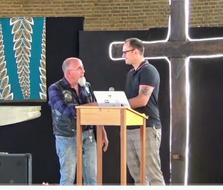Twee mannen staan naast een podium met een kruis en spreken met elkaar.