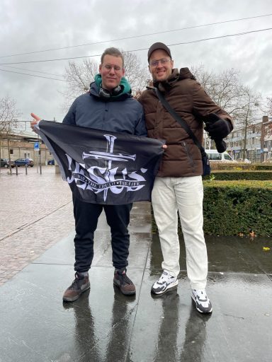 Twee mannen poseren met een vlag, buiten op een natte straat met bomen op de achtergrond.