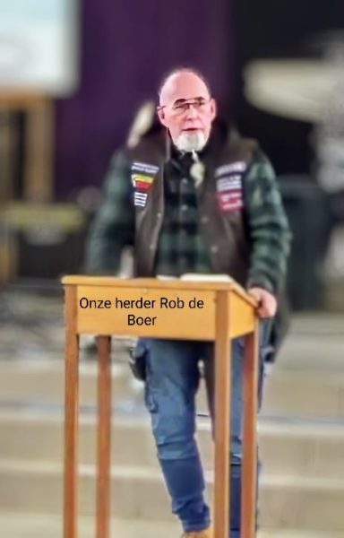 Man in een groen geruit overhemd en leren gilet spreekt achter een houten spreekgestoelte.